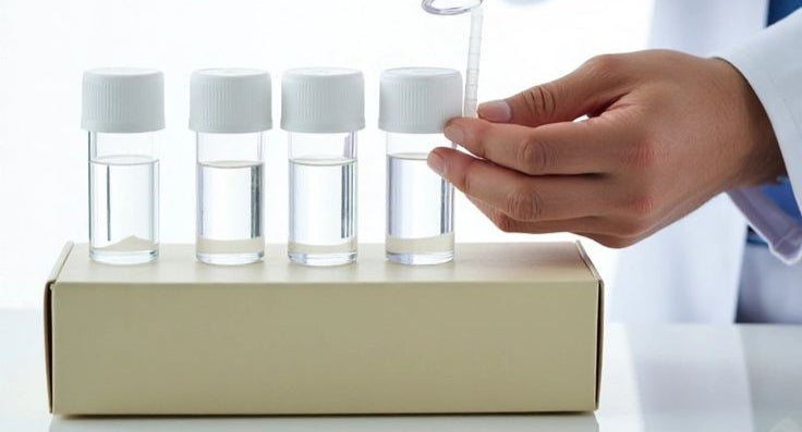 Person pouring liquid into small glass bottles with white caps on a beige box.